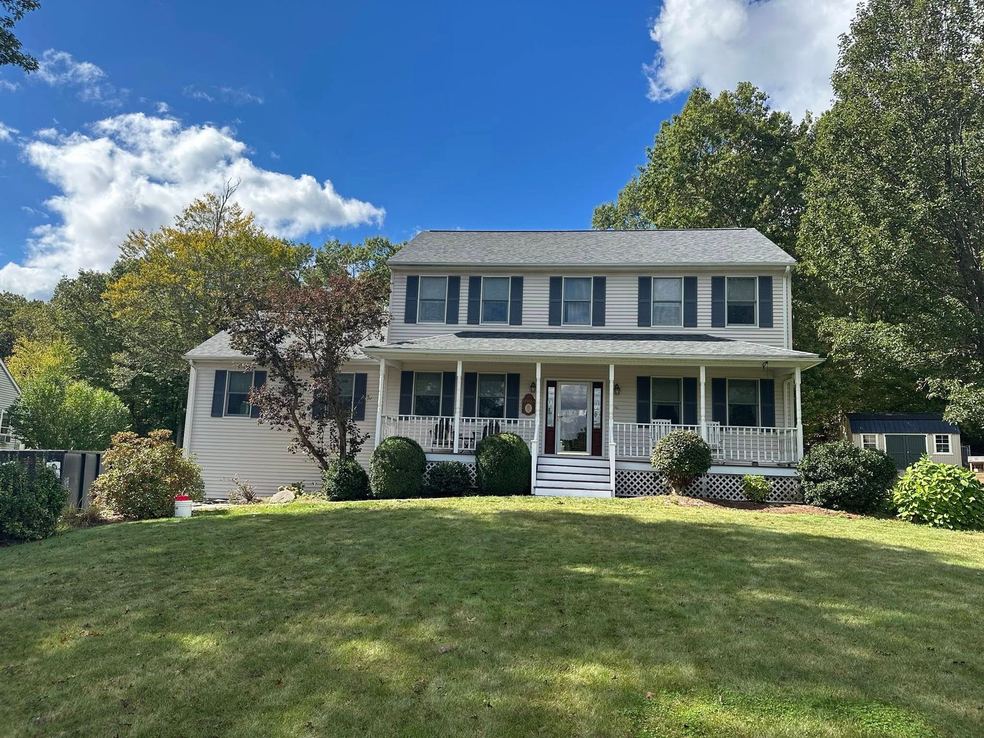Two-story beige house with porch, front lawn, trees, and a bright blue sky.