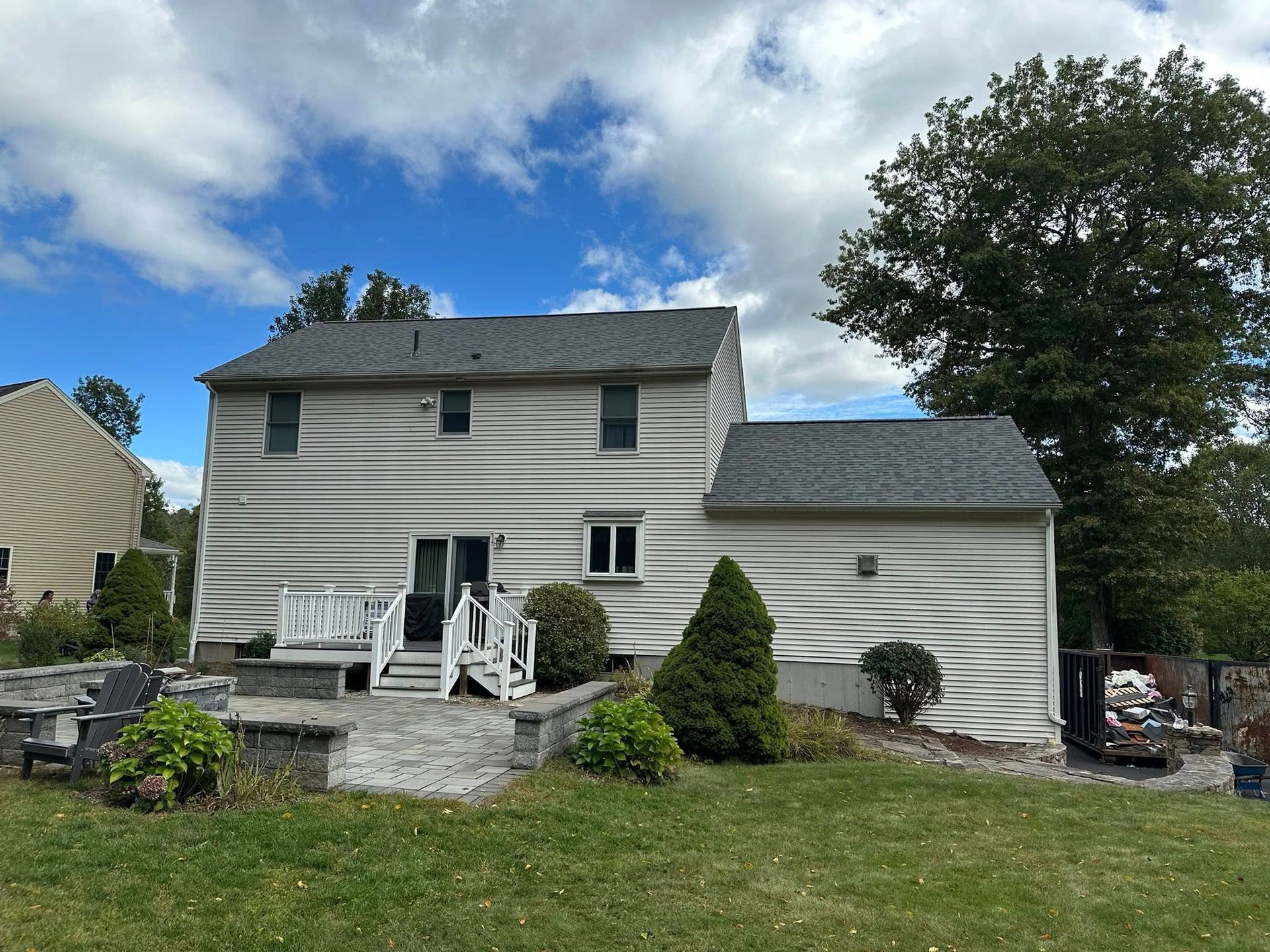 Back of a two-story house with a deck, stone patio, and green lawn under a partly cloudy sky.