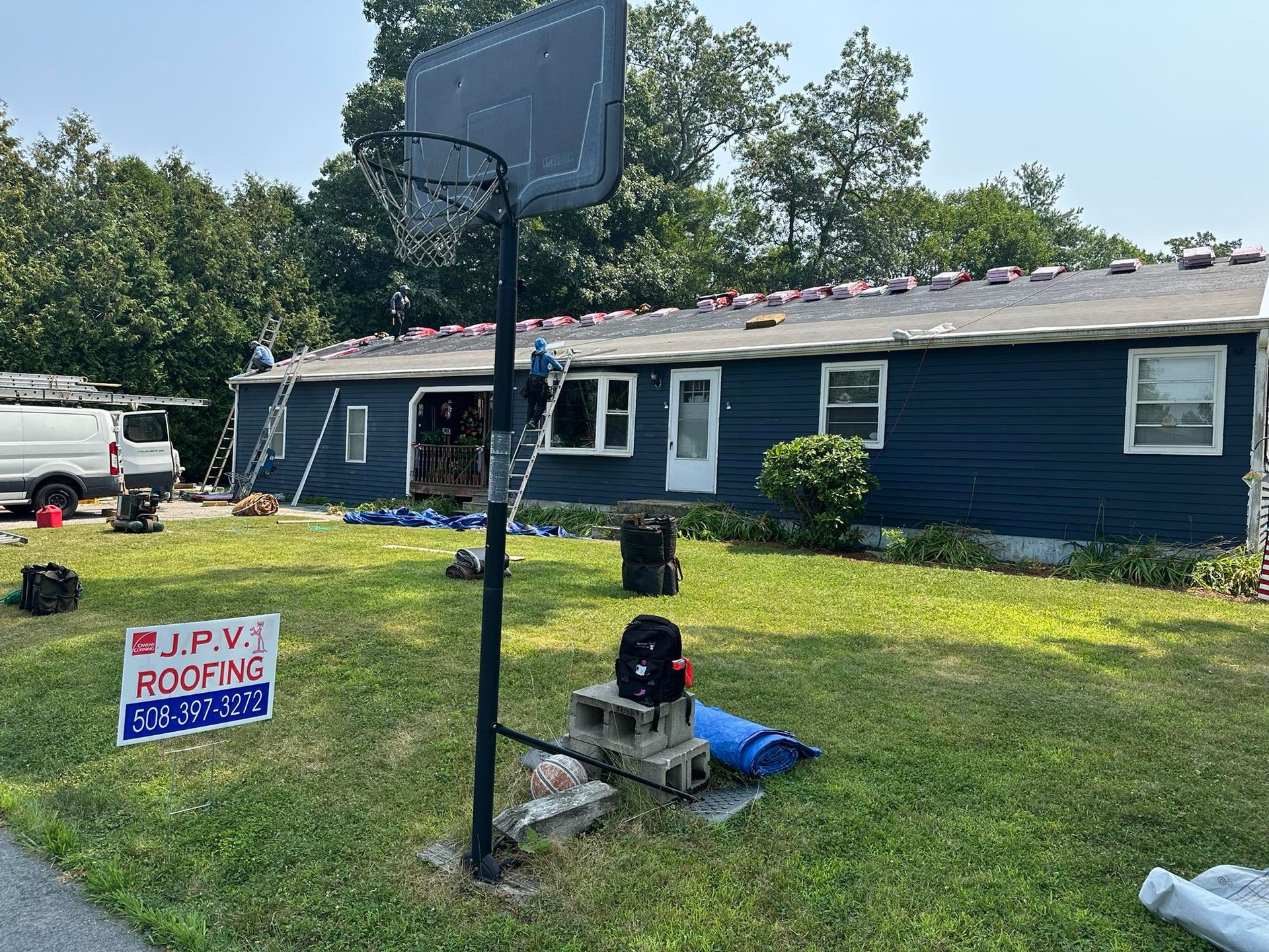 House roof replacement; blue house, basketball hoop, roofing crew working on roof, sunny day.