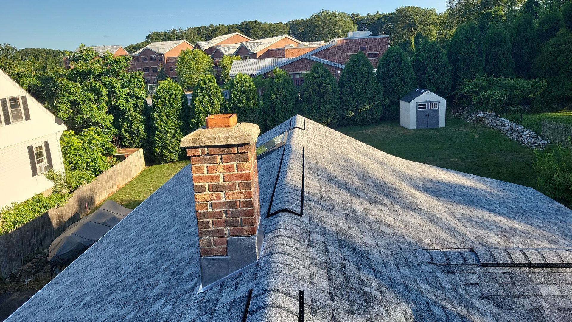 Roof with brick chimney, shingles, green trees, and buildings in the background on a sunny day.