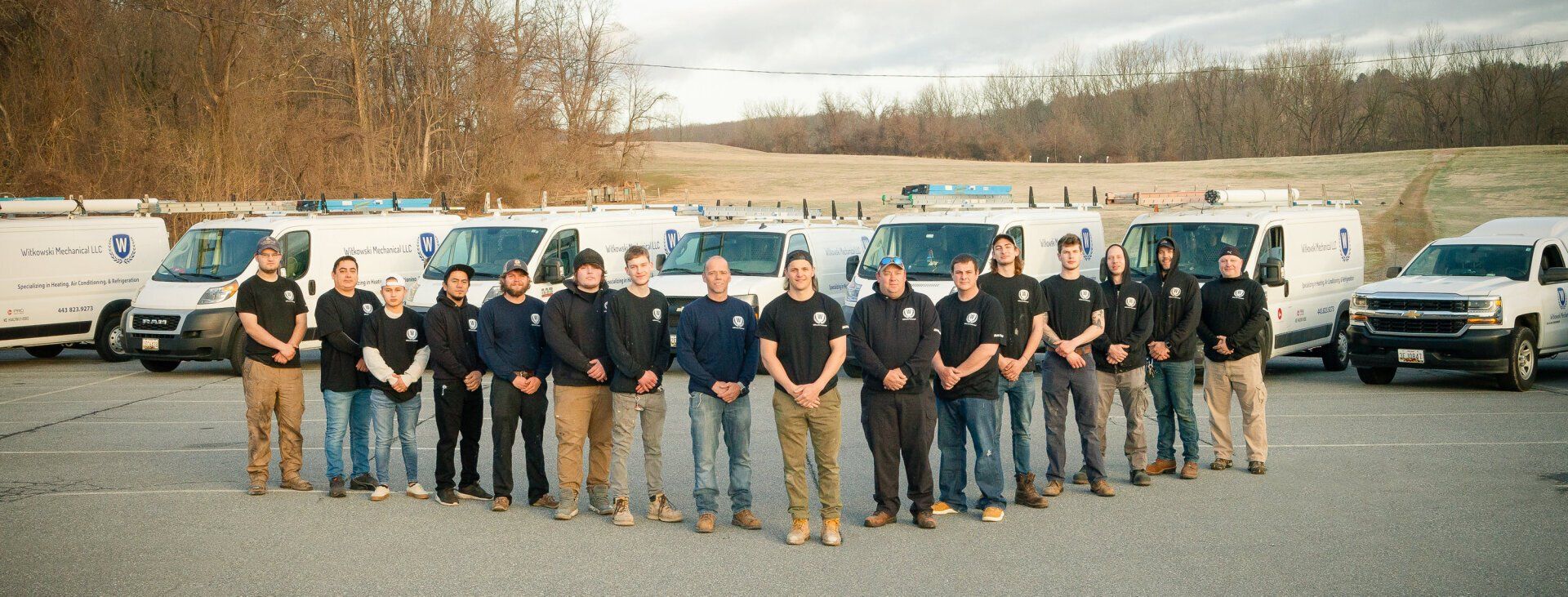 A group of men in work uniforms stand in front of several service vans and a field under a cloudy sky.