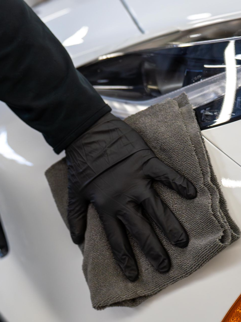 Gloved hand wiping a car surface with a gray microfiber cloth