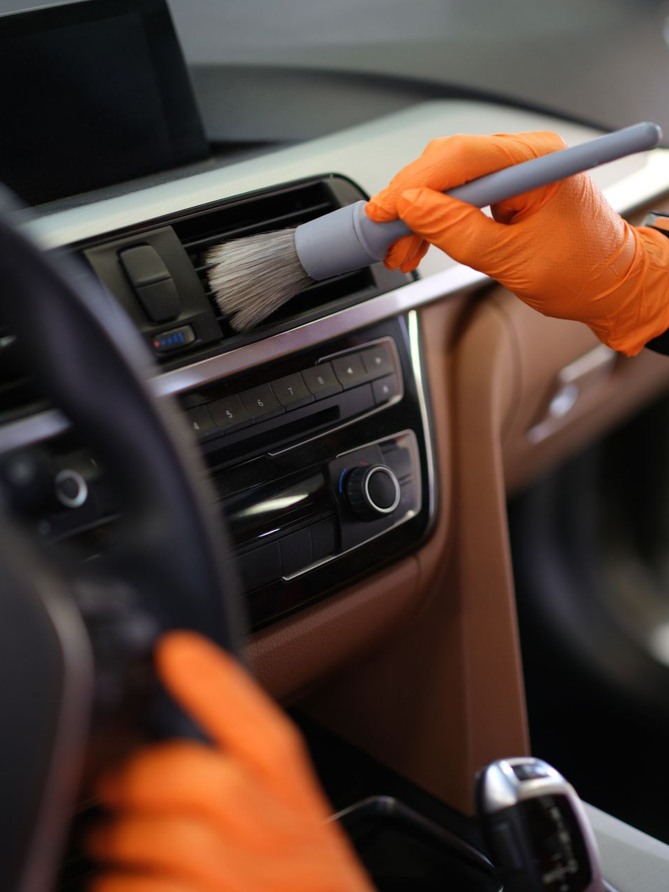 Orange-gloved hands cleaning a car dashboard vent with a brush and vacuum attachment