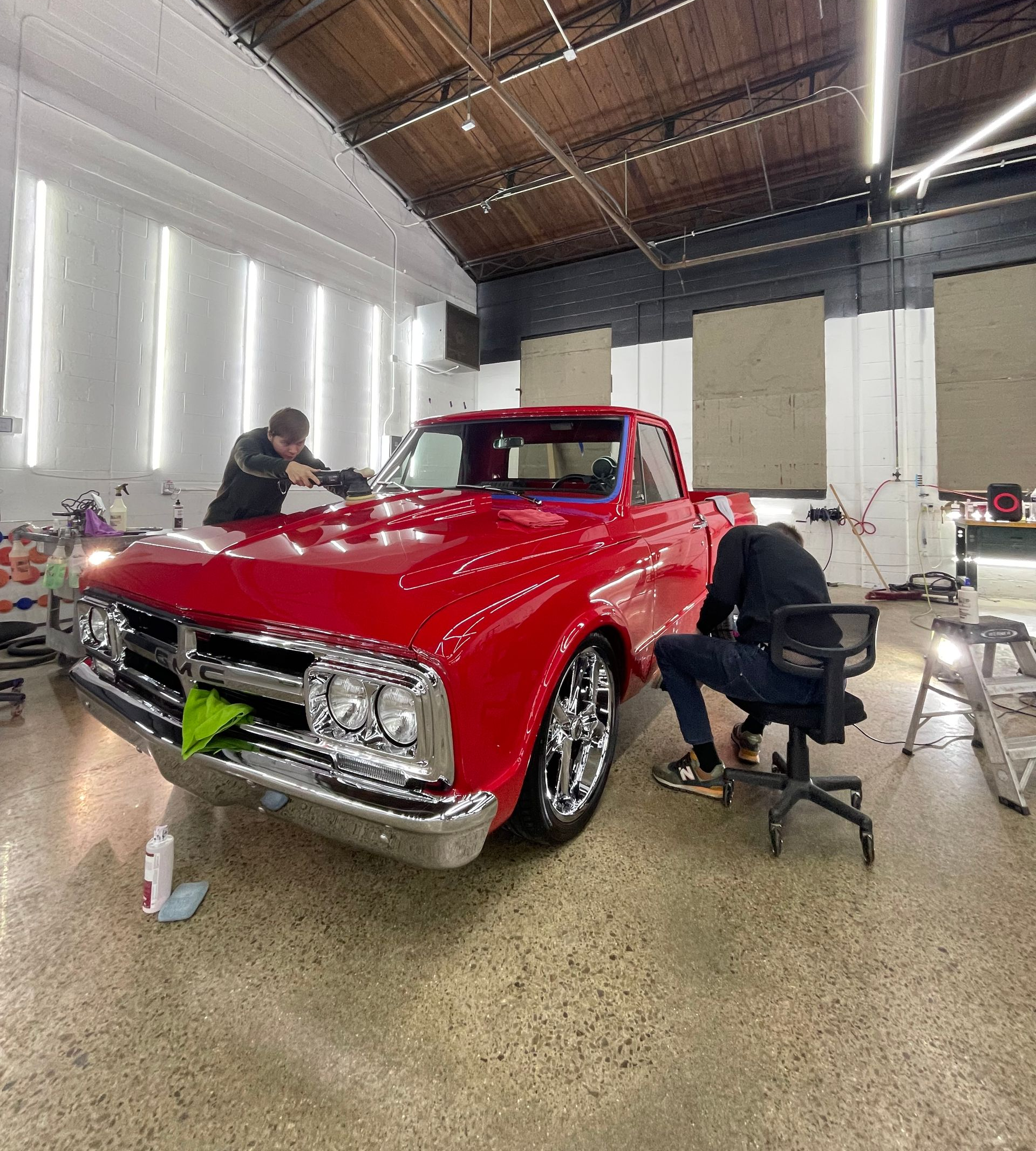 Red classic car being polished in a garage by two people, with other cars and stools nearby.
