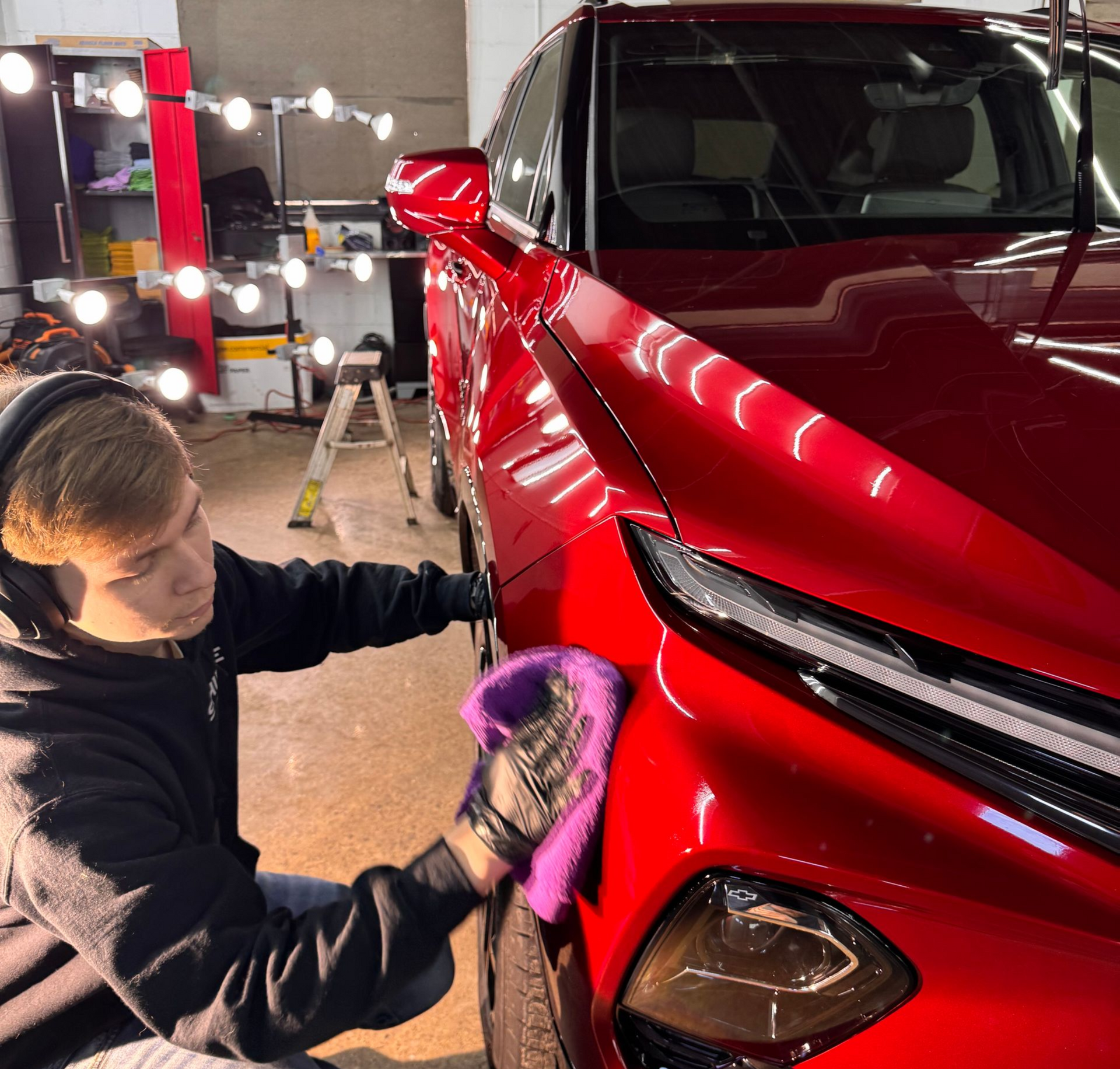 Person polishing a red car’s front fender in a garage with bright work lights.