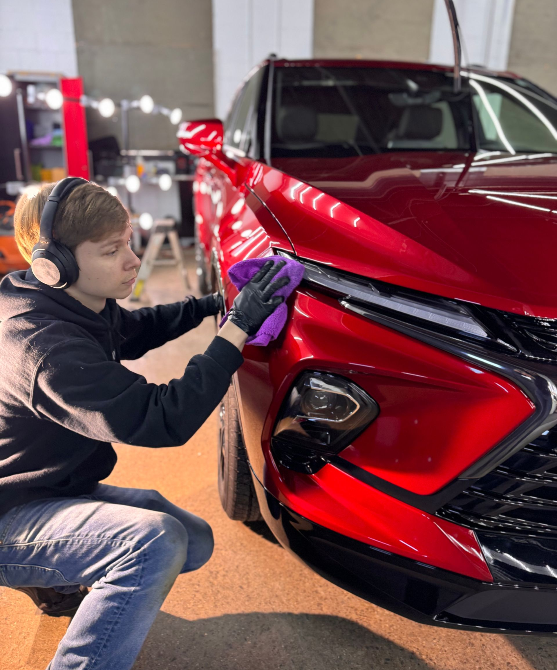 Person polishing the front of a red car with a purple cloth in a garage