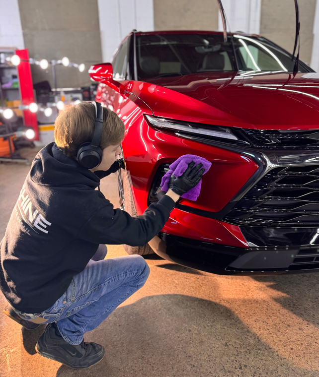 Person kneeling to detail a red car’s front bumper with a purple cloth in a garage