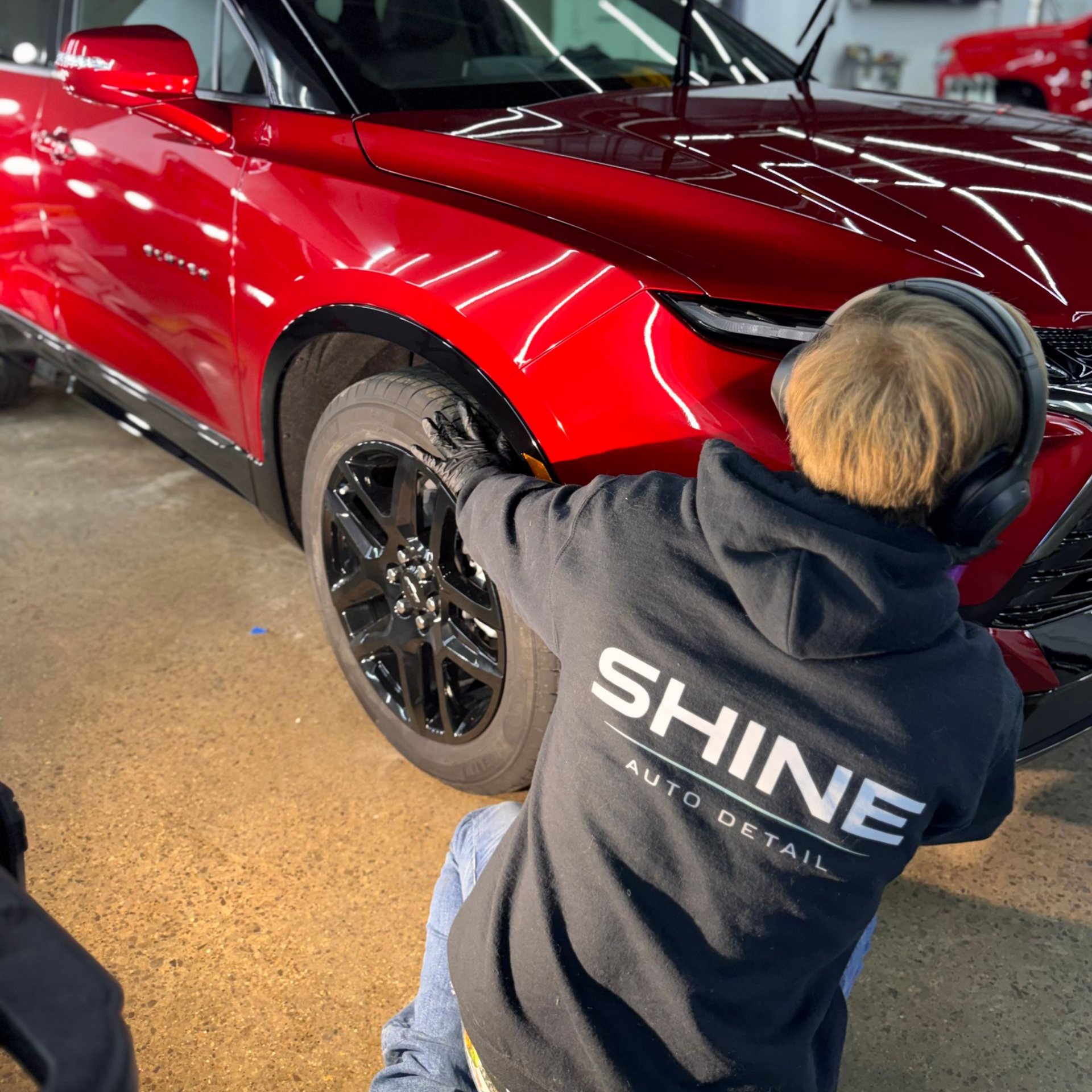 Person polishing the front wheel of a red sports car in a garage, wearing a SHINE hoodie