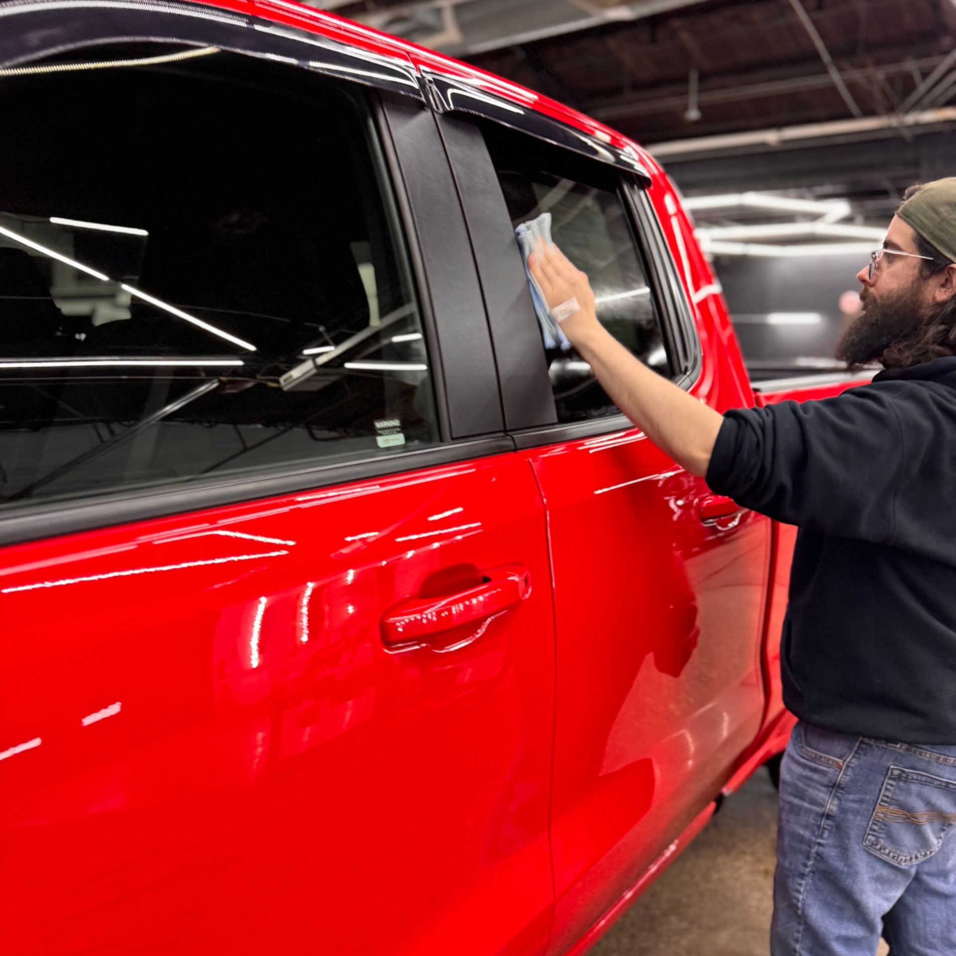 Man cleaning the rear window of a red pickup truck in a garage