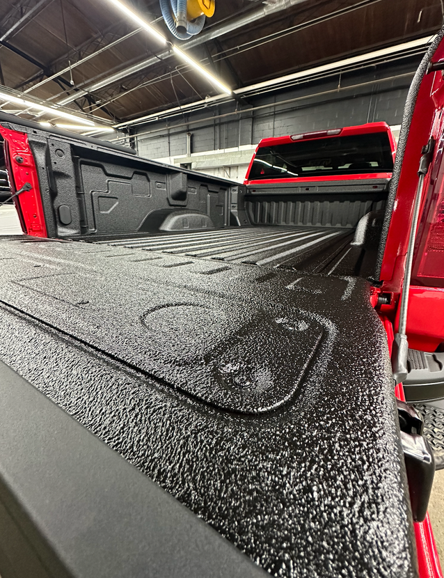 Red pickup truck bed with black textured bed cover inside a garage or workshop.