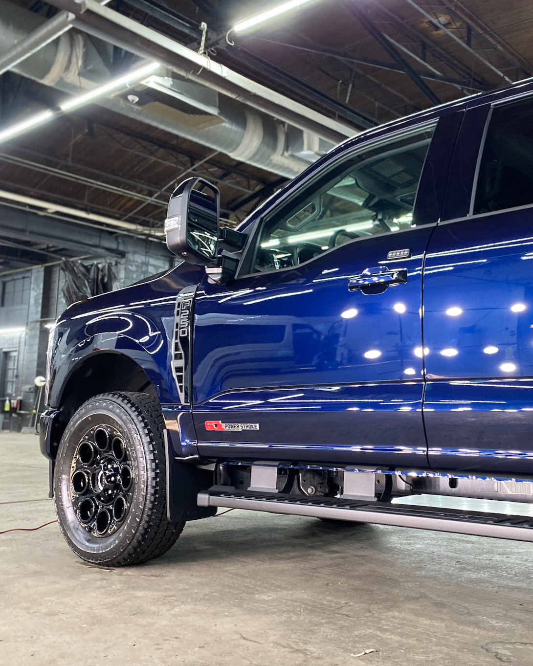 Blue lifted pickup truck in an indoor garage, shown from the front side with black wheels and steps.