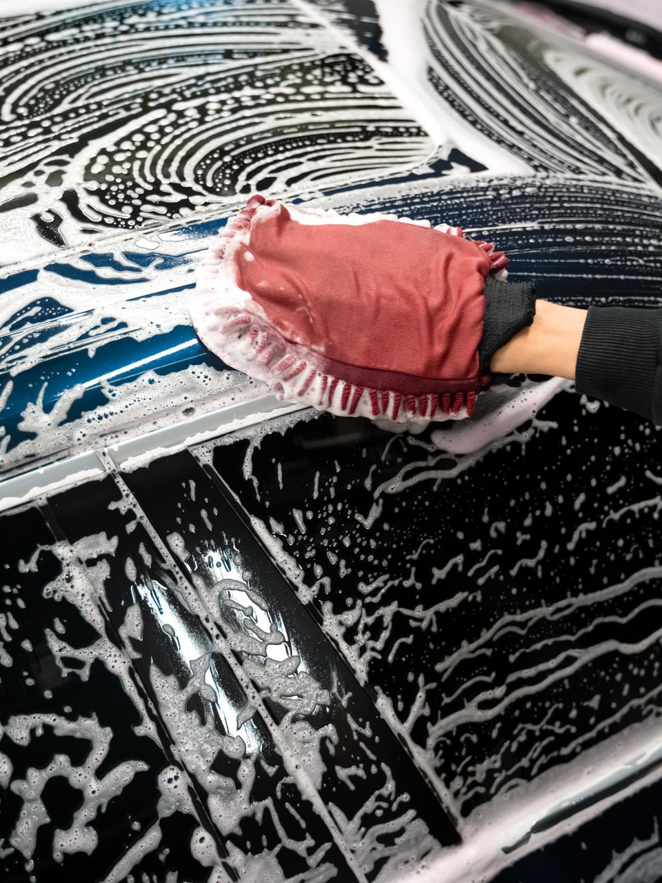 Gloved hand washing a black car with pink sponge and white soap suds