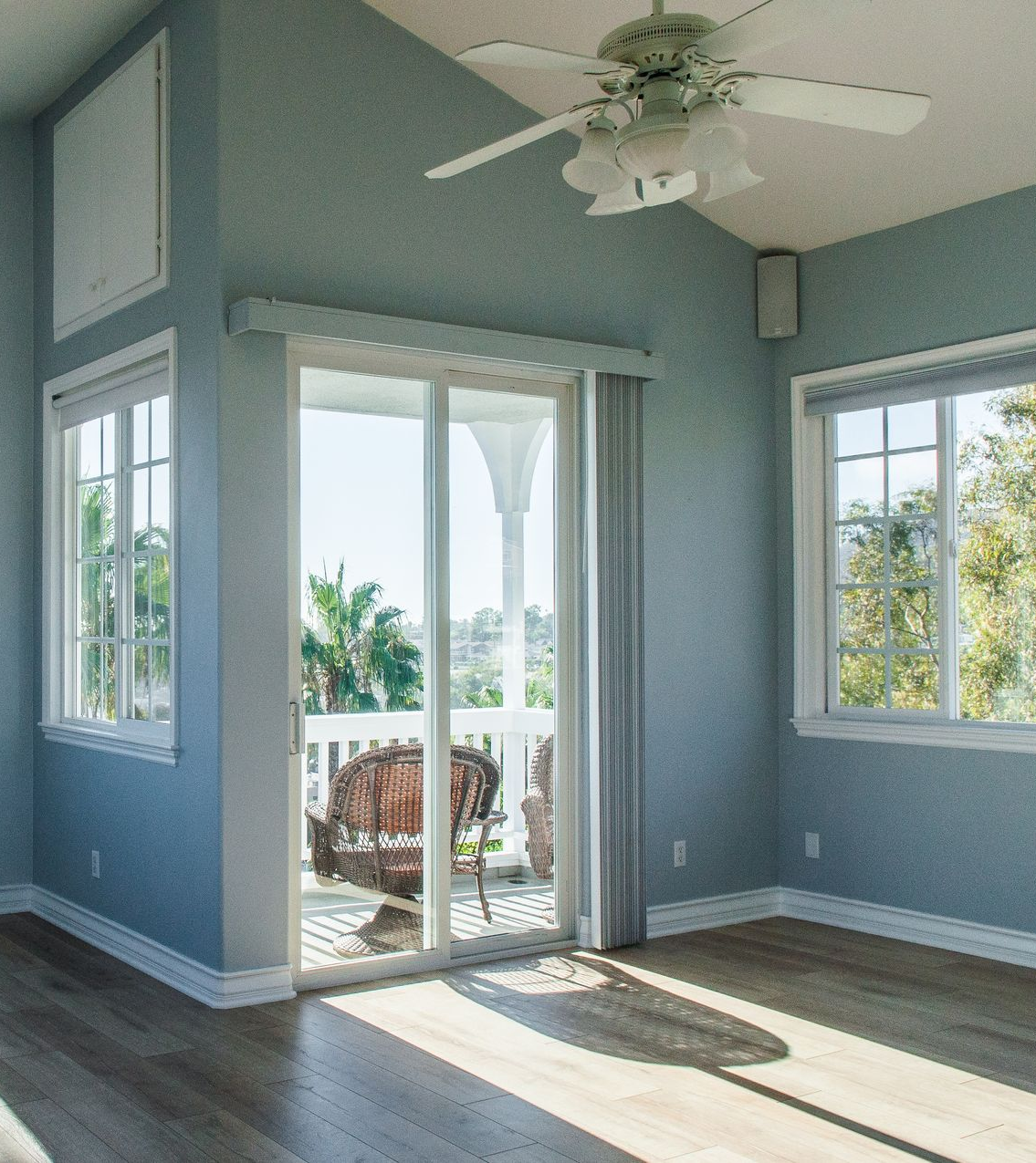 Empty sunlit room with light blue walls, white ceiling fan, and sliding glass doors to a patio.