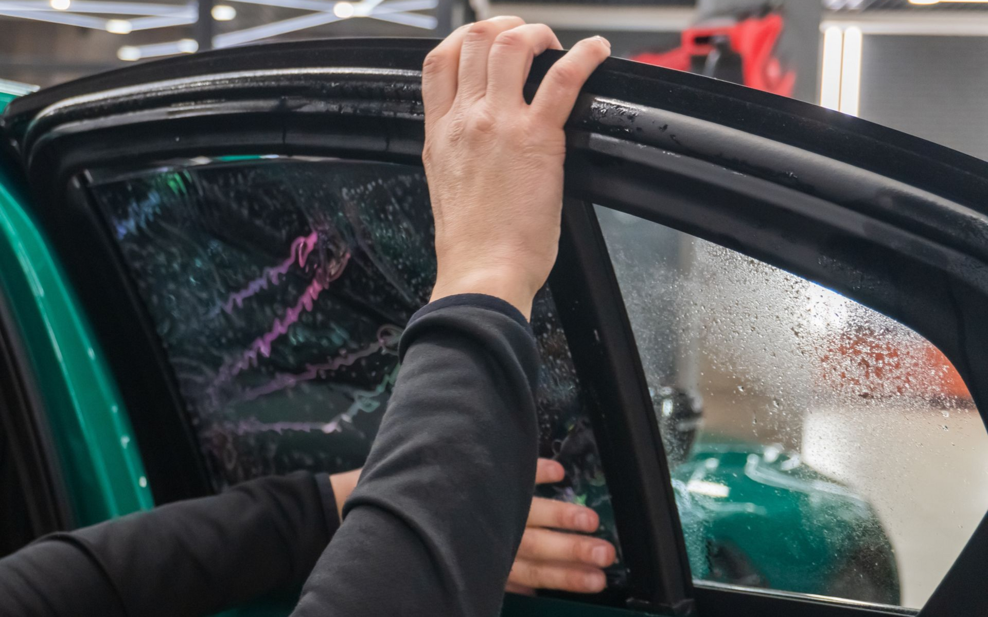 Hands opening a black car door in a rainy garage, with a person visible inside