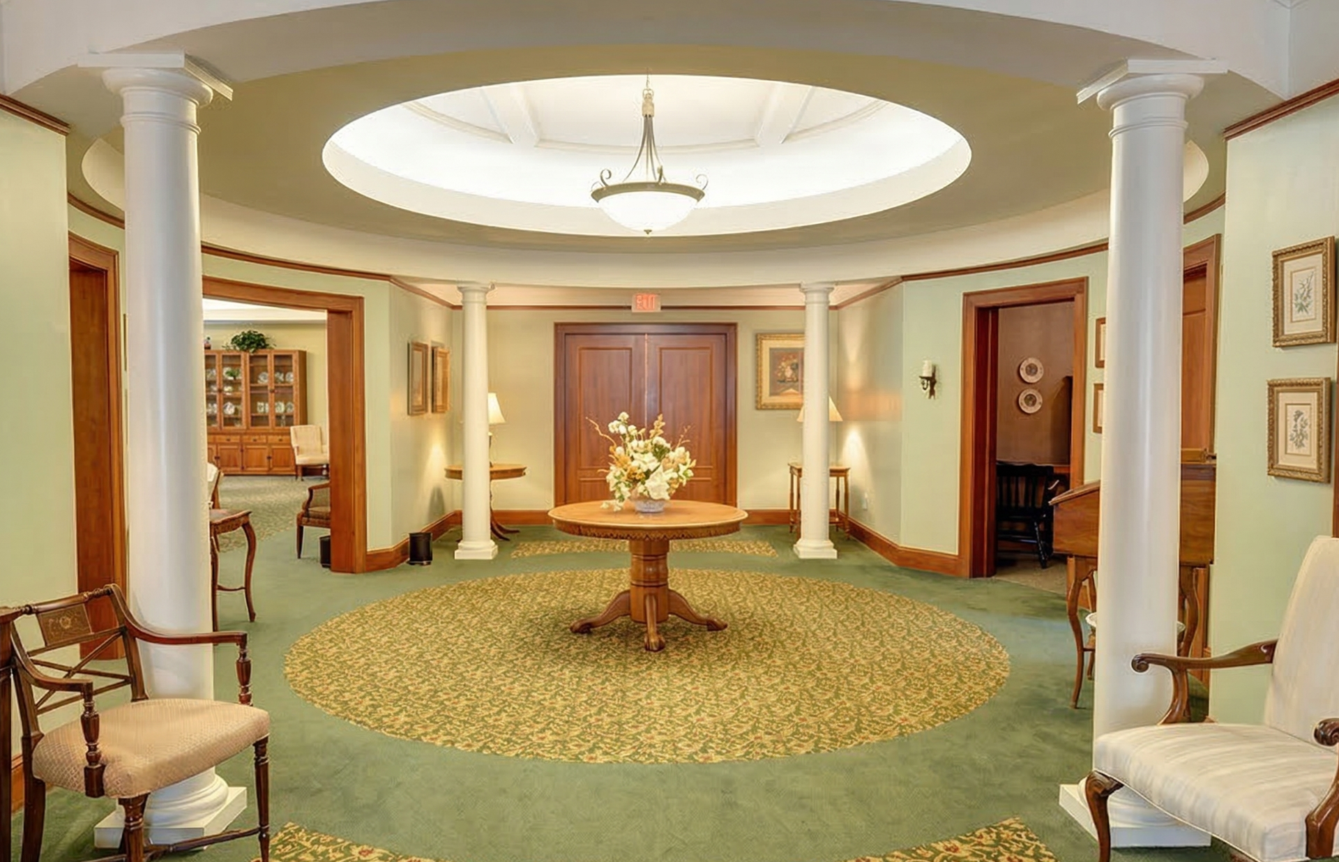 Elegant hotel lobby with round skylight, central floral table, and white columns against green carpet.