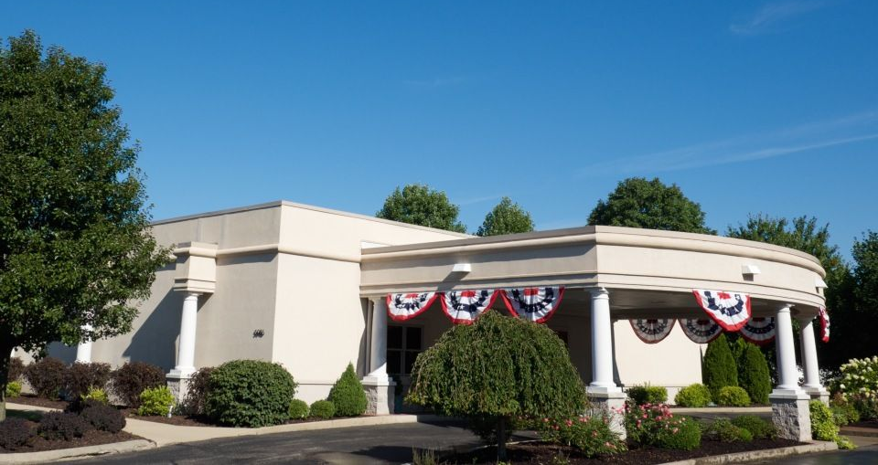 A curved, stone-pillared building entrance decorated with patriotic red, white, and blue bunting under leafy trees.