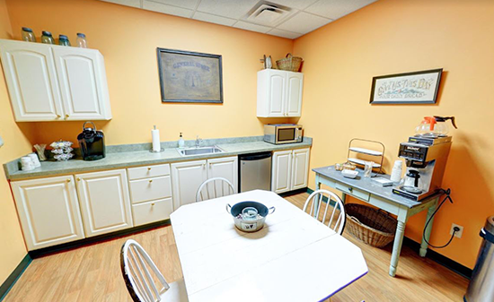A kitchen break room with peach walls, white cabinets, a small dining table with chairs, and a coffee machine.