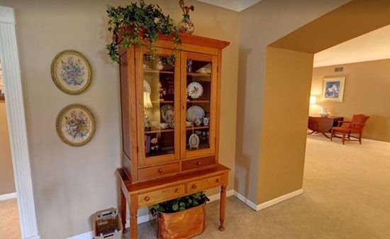 A wooden hutch with glass doors stands against a wall with two floral plates, overlooking an open room with furniture.