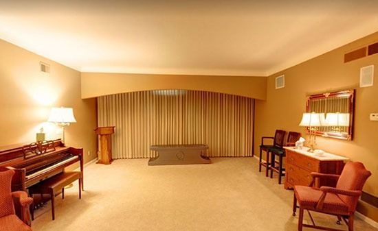 A spacious, warm-toned room with a piano, a wooden lectern, a bench, two chairs, and a mirror on the wall.
