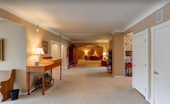 A spacious hallway with beige carpet and walls, leading to a living area with a small wooden desk in the foreground.