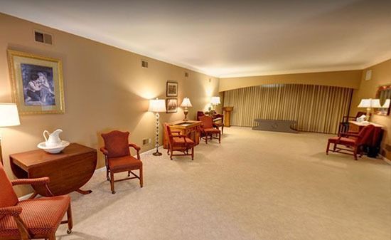 A spacious, beige-carpeted room furnished with several red armchairs, small wooden side tables, and warm wall lighting.