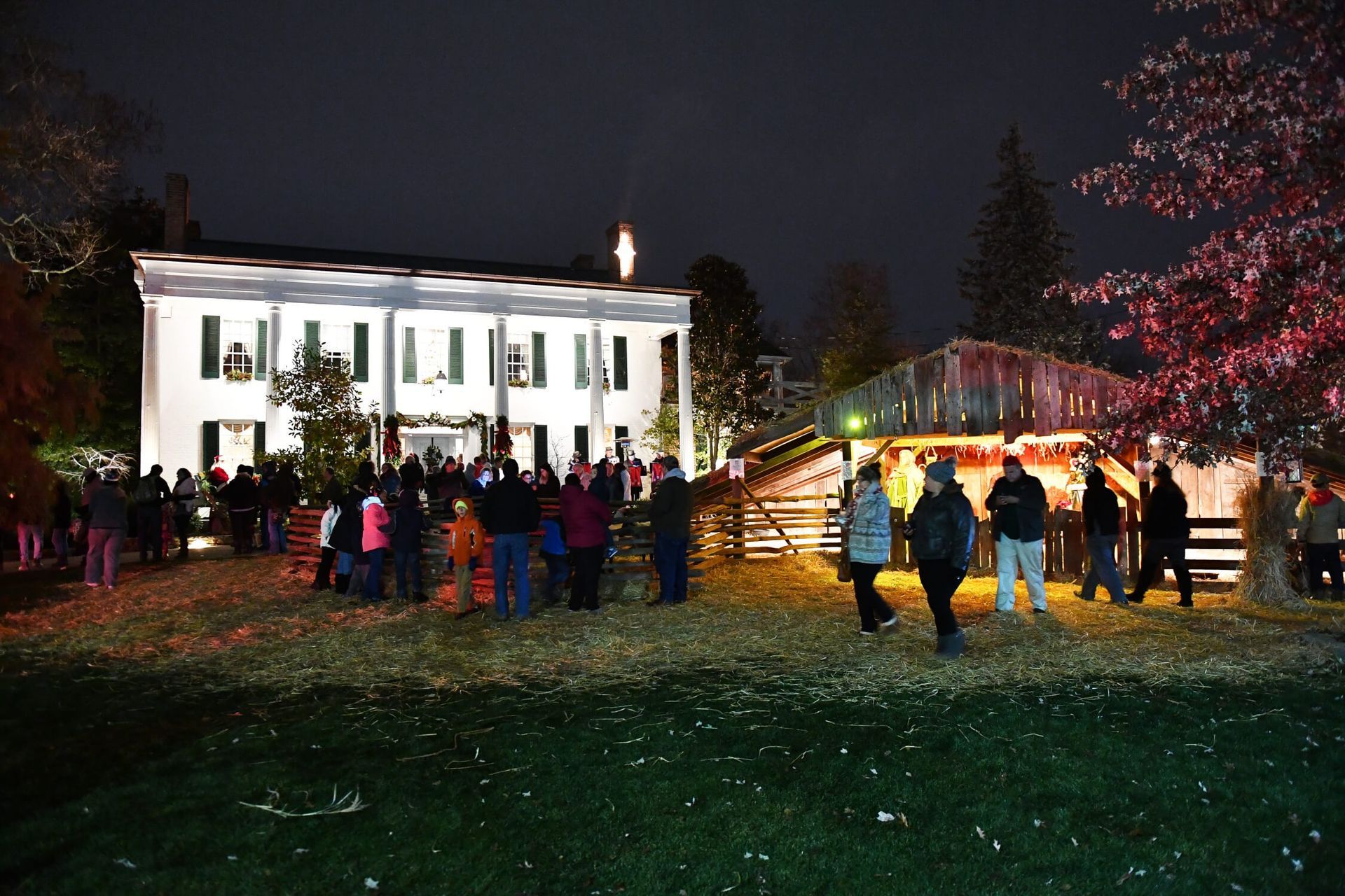 A group of people gather at night outside a white, illuminated historic house and a wooden structure during an event.