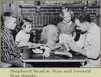 A man shows three children a wax sculpture of a head and hands on a table in front of a brick fireplace.