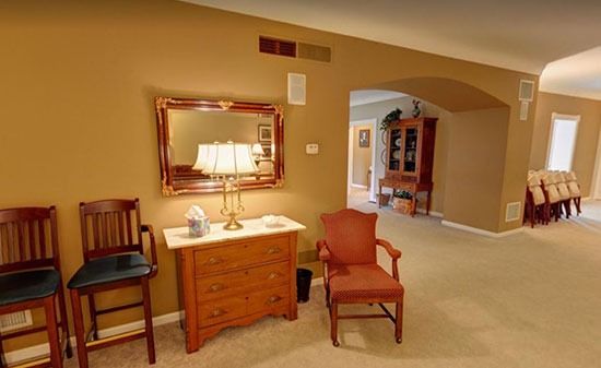 A room with two wooden chairs, a chest with a lamp and mirror, and an armchair, leading to an arched hallway entrance.