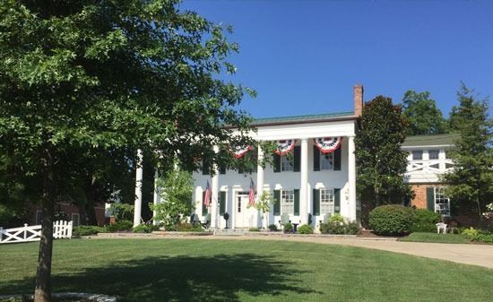 A large, white, two-story house with tall pillars, decorated with American bunting, surrounded by a lawn and trees.