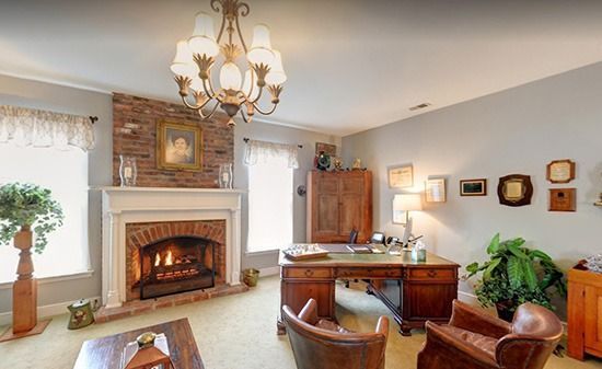 Home office with a lit fireplace, wood desk, two leather chairs, a chandelier, and decor on walls.