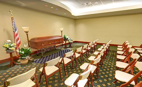 A funeral parlor viewing room with rows of chairs facing a wooden casket, an American flag, and floral arrangements.