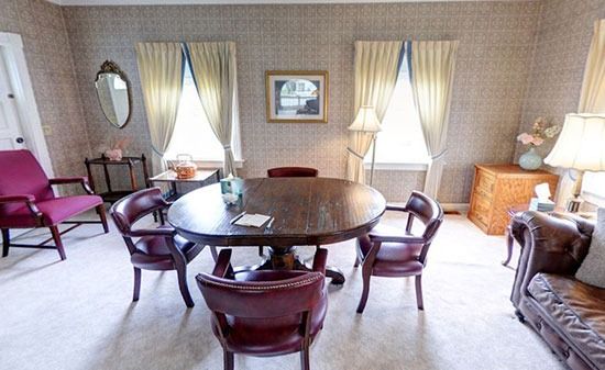 A room with patterned wallpaper, a circular wooden table with four burgundy chairs, a side armchair, and a sofa.
