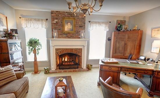A study with a lit fireplace, brick chimney breast, chandelier, large wooden desk, and comfortable seating.