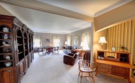 A living room with a large wooden bookshelf, light-colored carpet, a dining area, and a desk with a lamp.