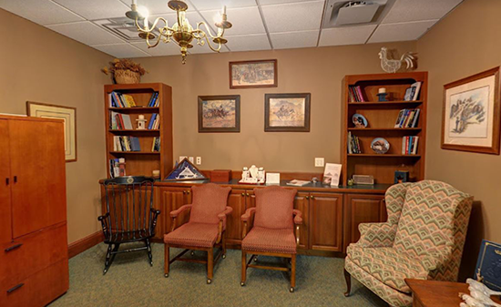 A room with two bookshelves, two red chairs, and a patterned armchair, set against tan walls with framed artwork.