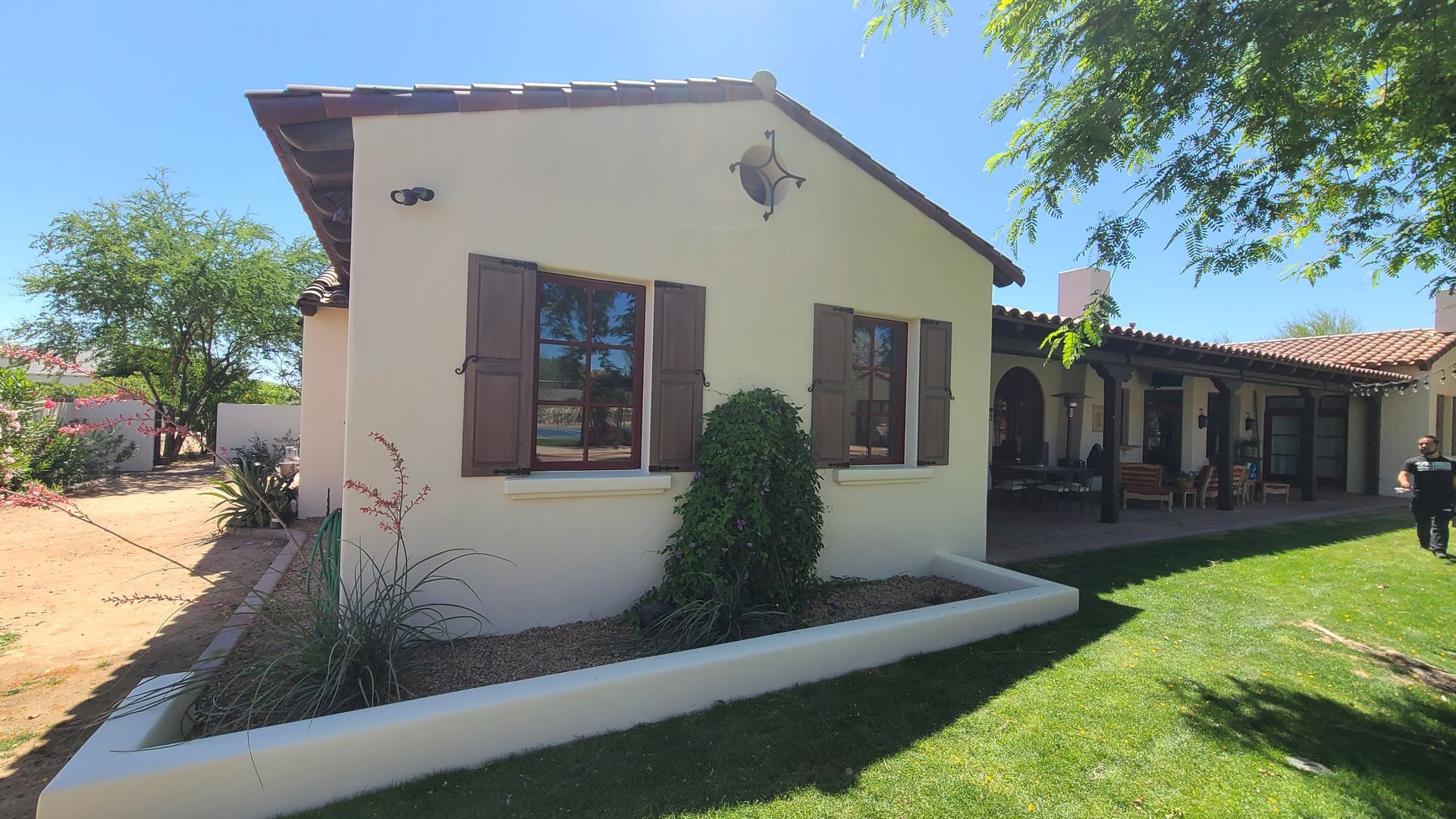 Tan stucco house with brown shutters, a tiled roof, and a covered patio on a sunny day.
