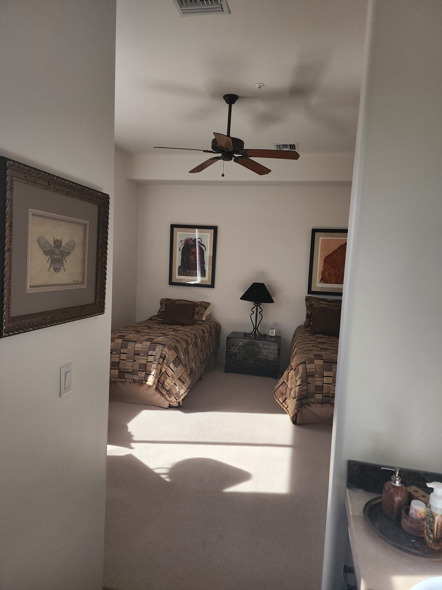 View of a bedroom with two twin beds, art, and a ceiling fan, seen through a doorway.