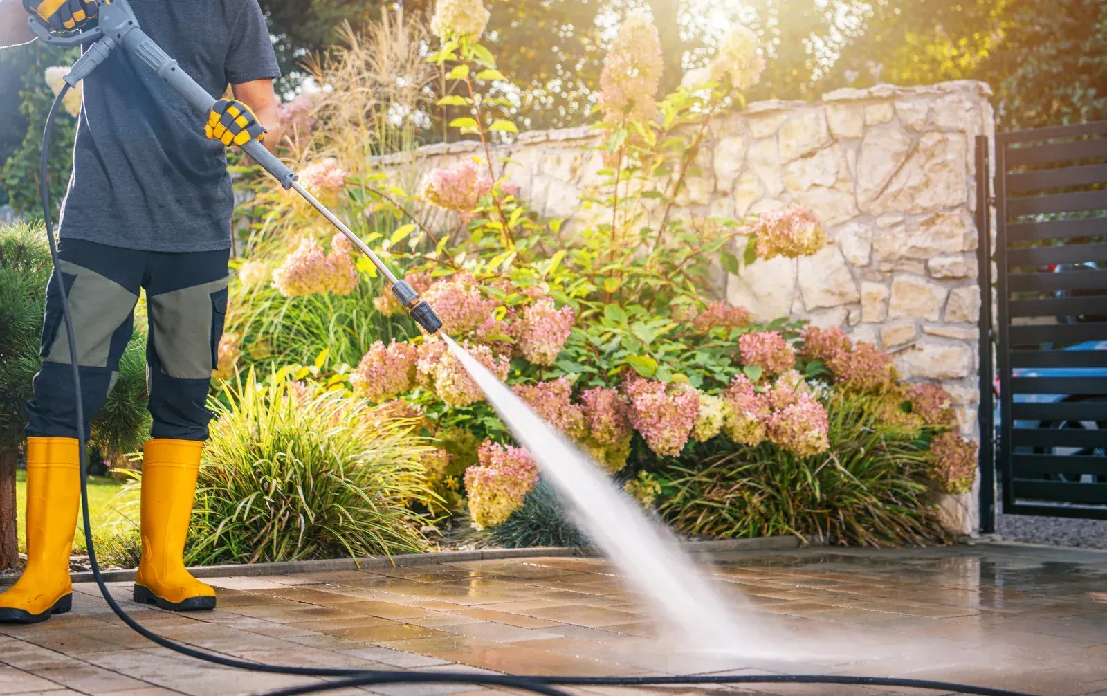 Person power washing a patio; yellow boots, water spray, garden in background.