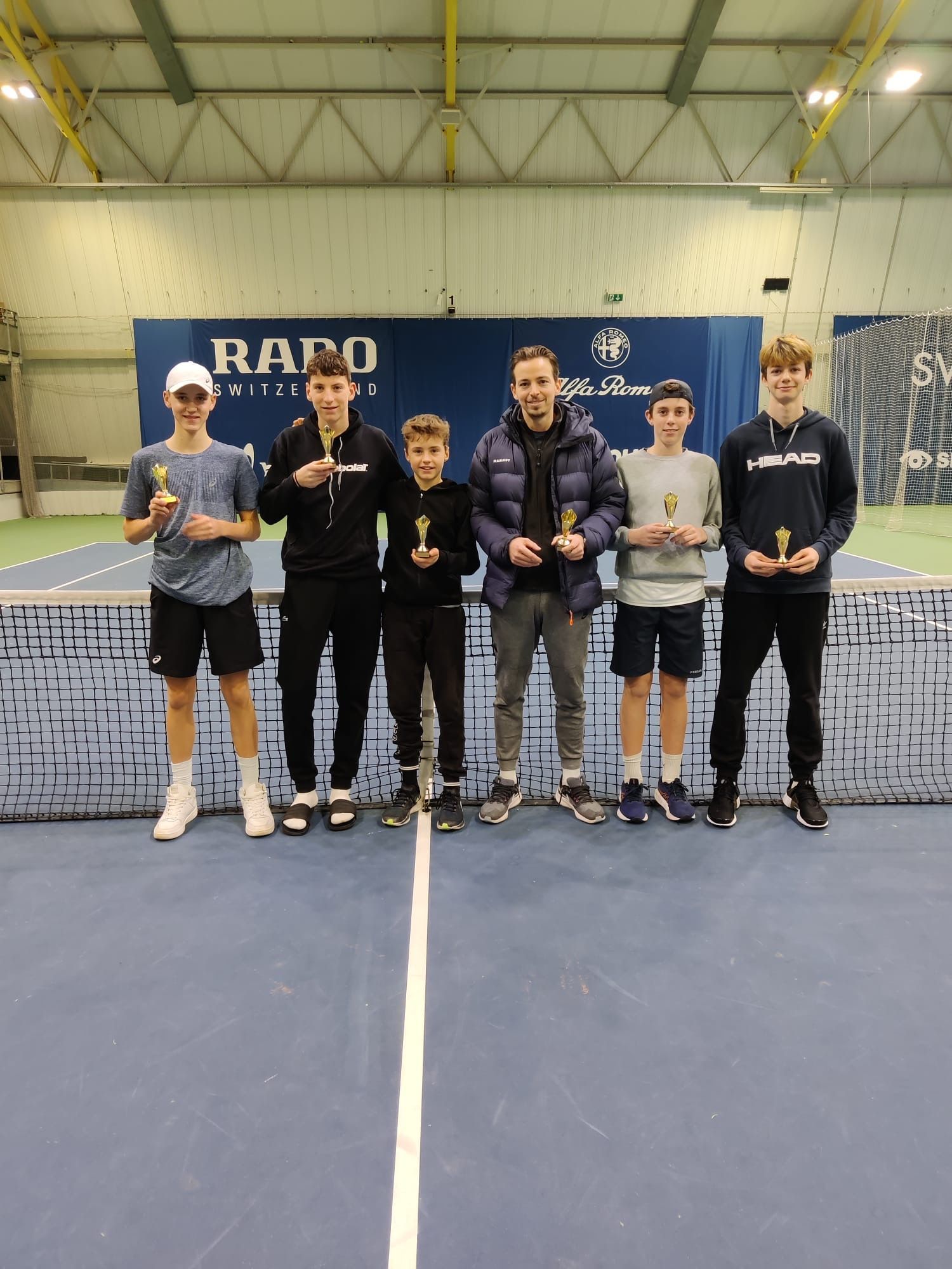 Eine Gruppe junger Männer steht mit Trophäen in den Händen auf einem Tennisplatz.