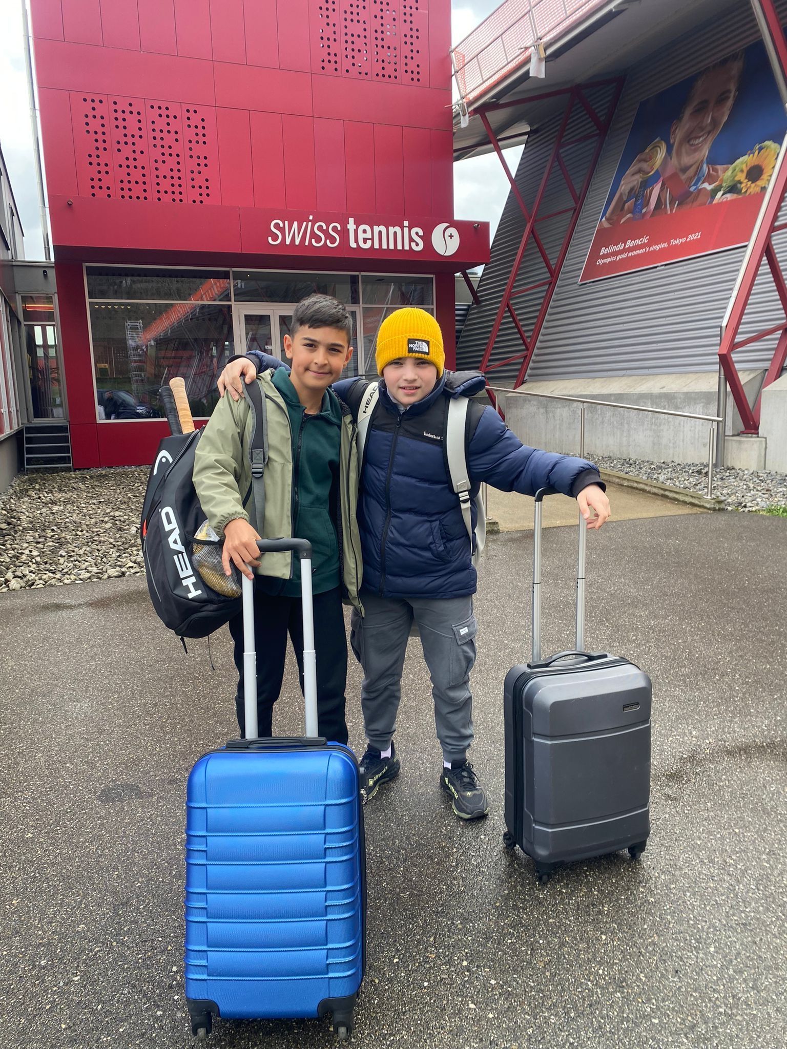Zwei kleine Jungen stehen mit Gepäck nebeneinander vor einem Gebäude.
