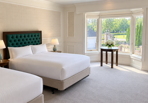 Bright hotel bedroom with white bedding, teal headboard, and a bay window overlooking a green garden