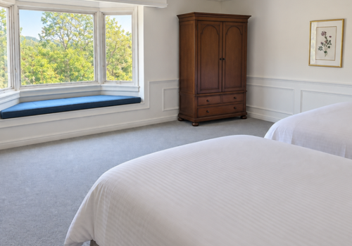 Bright bedroom with large window seat, wooden armoire, and two white beds