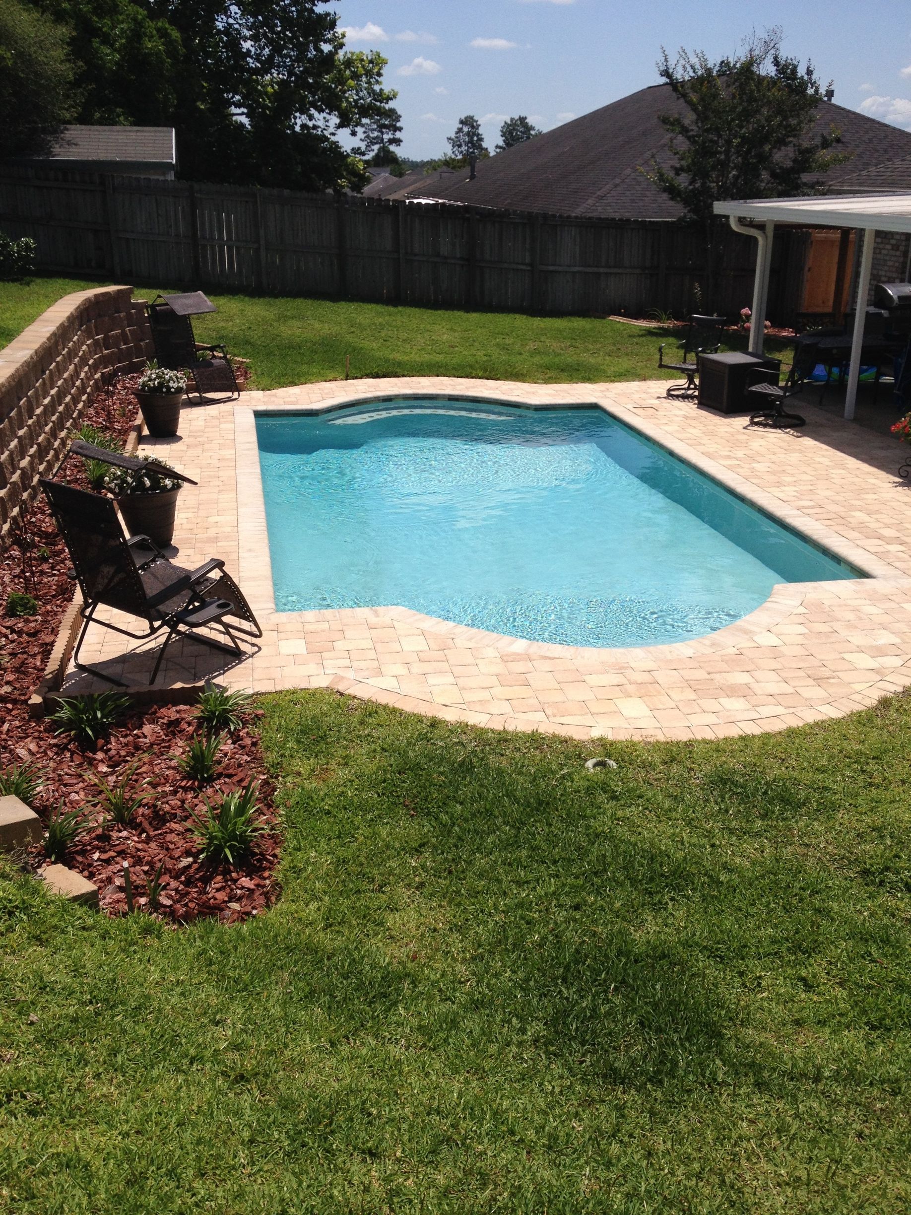 A backyard swimming pool with light blue water and stone pavers, bordered by a brick retaining wall and a grassy lawn.