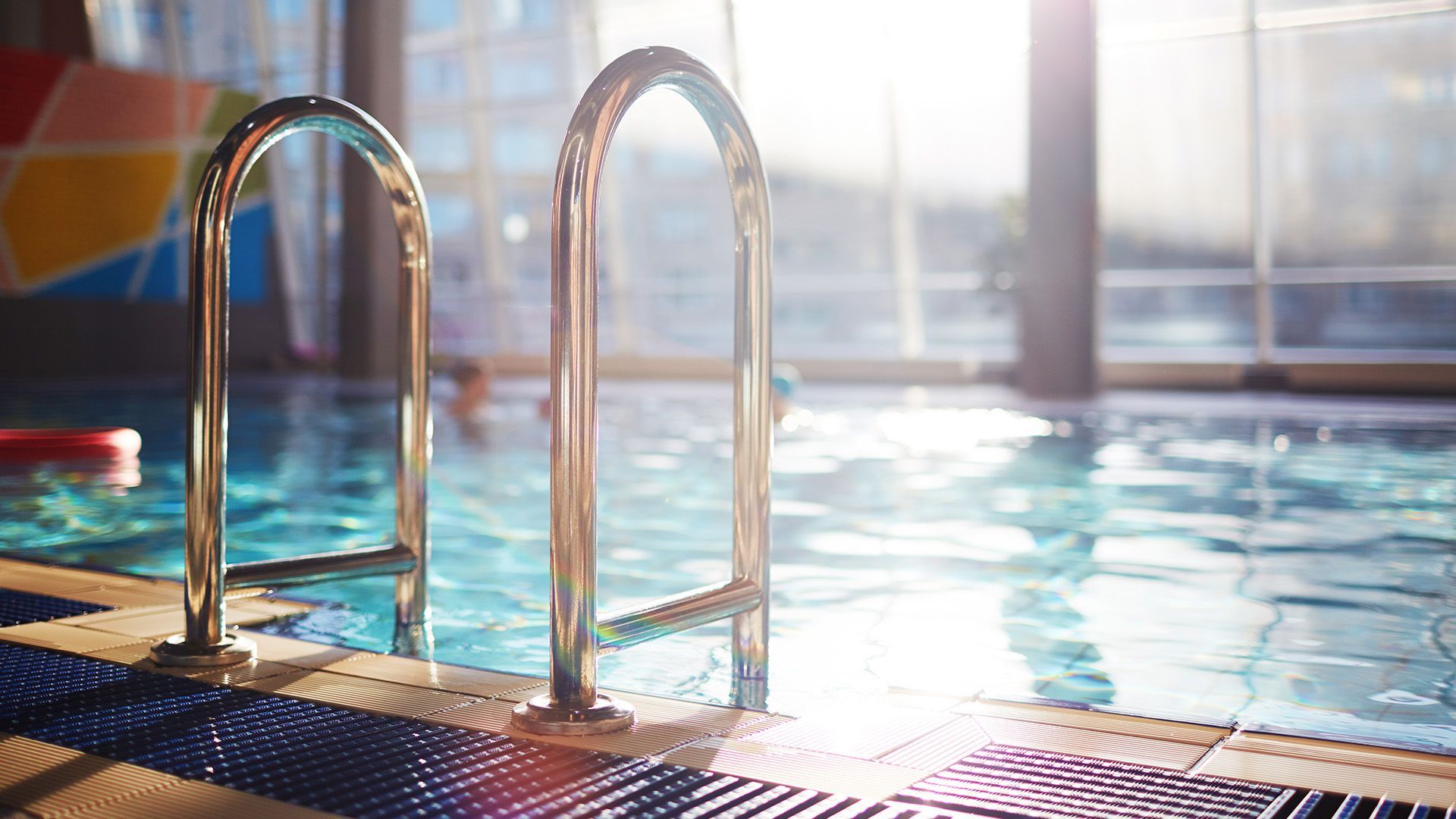 Two shiny metal pool ladders stand at the edge of an indoor swimming pool bathed in bright sunlight.