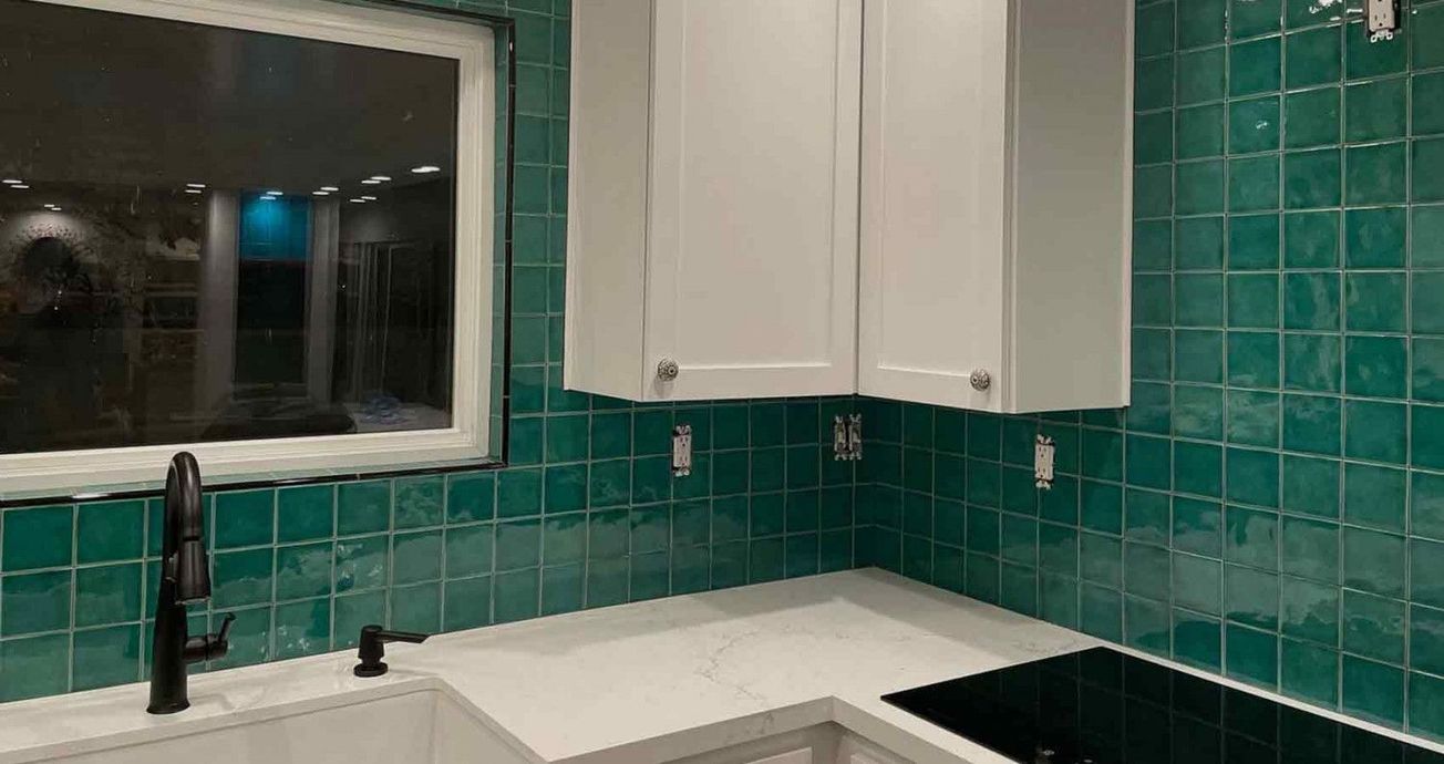 Modern kitchen corner with teal tile backsplash, white cabinets, black faucet, and sink by a window