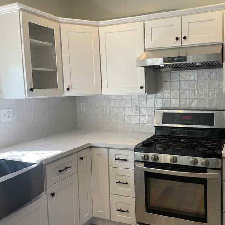 Bright white L-shaped kitchen with white cabinets, tiled backsplash, and stainless steel stove and sink