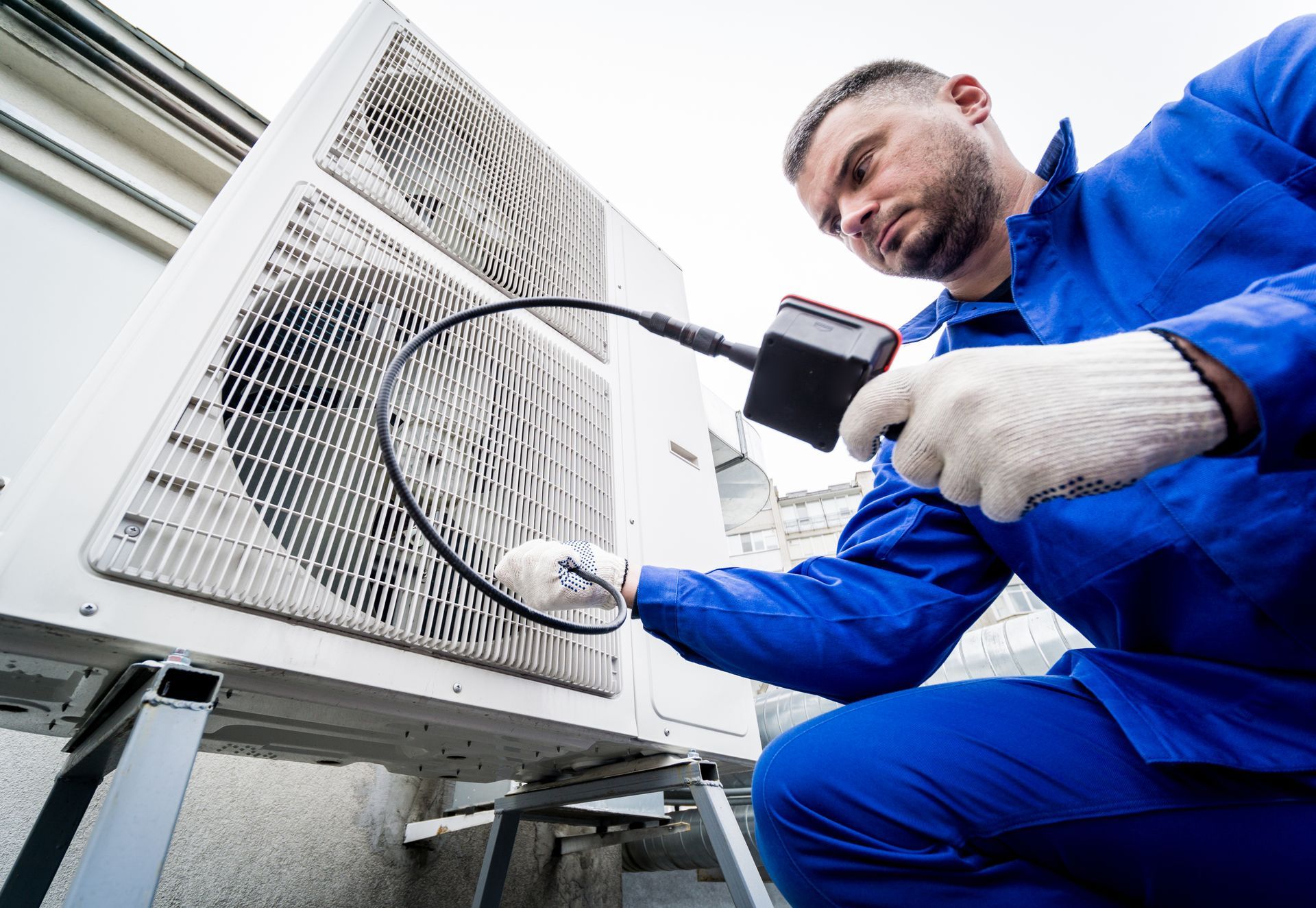 Man in blue overalls inspects an air conditioning unit with a tool.