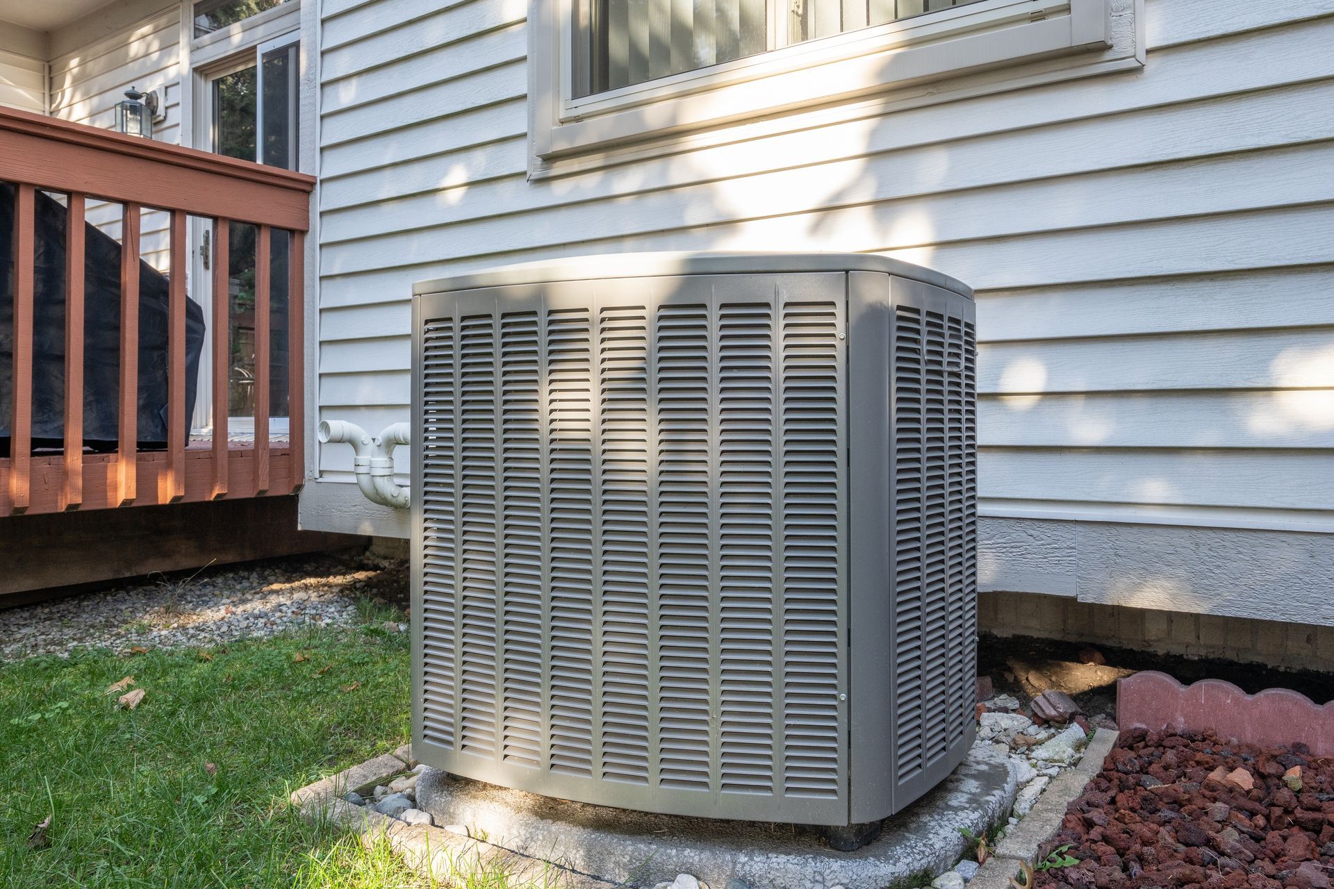 Air conditioning unit against a white house wall, near a deck, on a concrete pad.