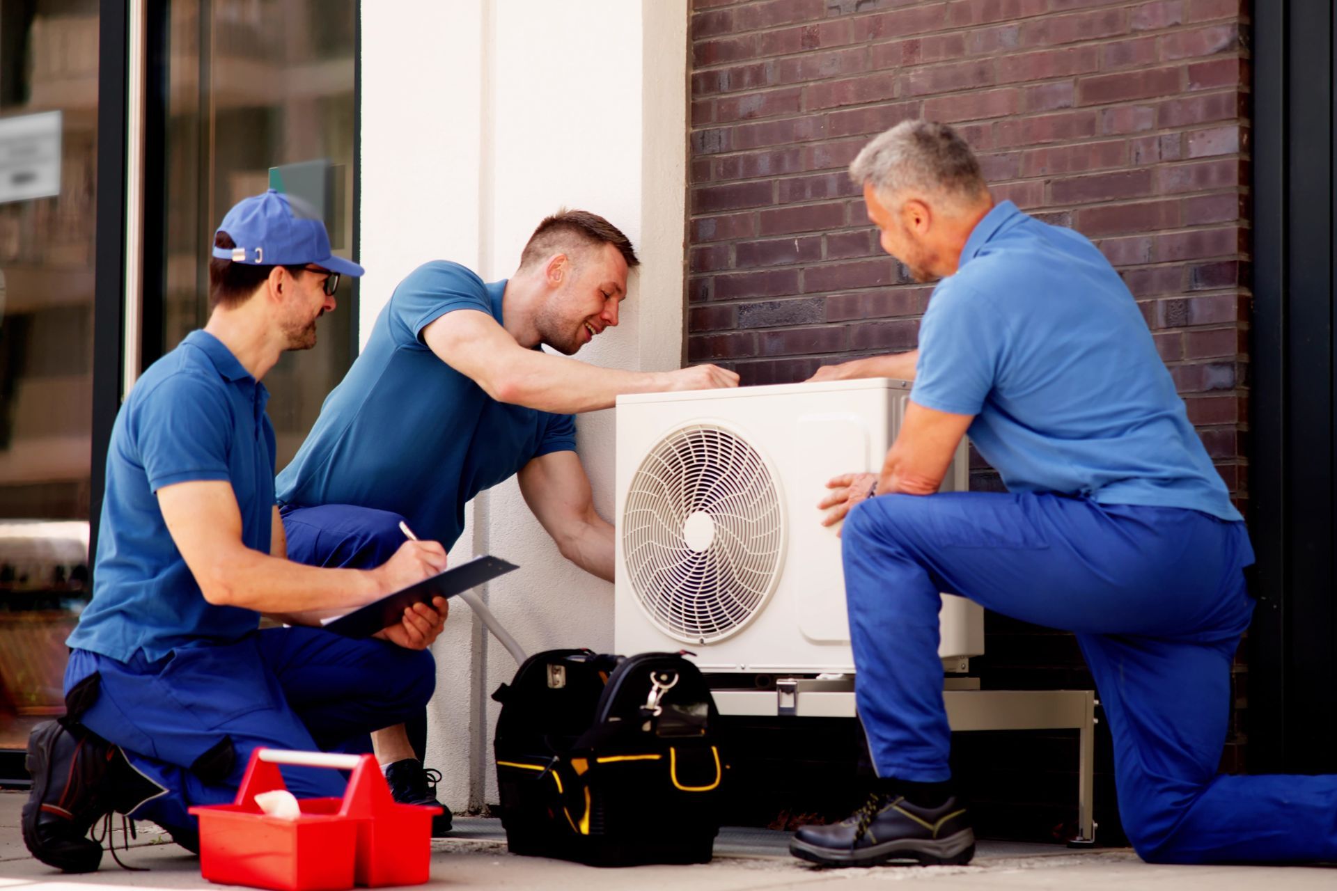 Three technicians in blue uniforms examine an outdoor HVAC unit, one kneeling with a clipboard.