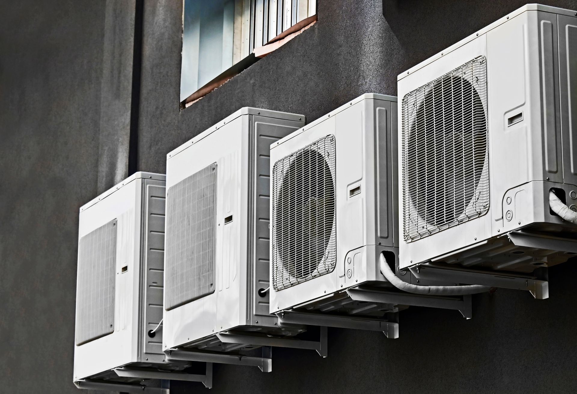 Four white air conditioning units mounted on a dark gray wall beneath a window.